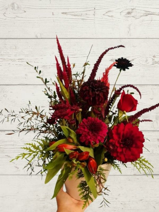 A red, brown, and green floral arrangement with various flowers including shades of red and a few green leaves, held against a white wooden background.