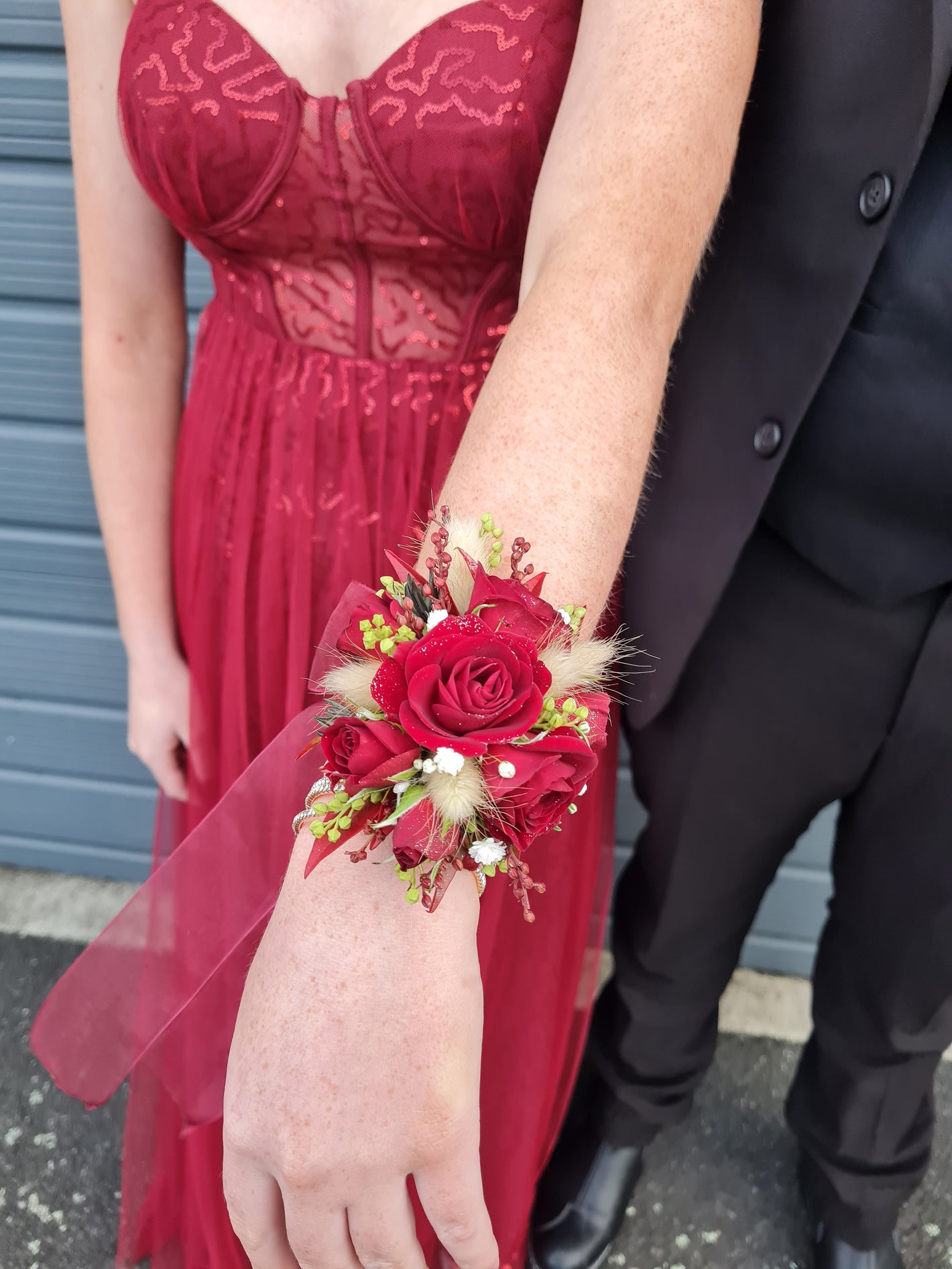 A close-up of a wrist corsage featuring red roses, green leaves, and white berries worn on a person's wrist, with a red lace dress and a black suit jacket visible in the background.