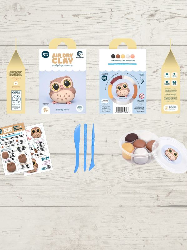 Owl-themed air dry clay kit laid out on a light wood background, including two retail boxes, three blue sculpting tools, instruction sheets, and a small container of mixed clay colors.