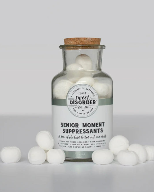 A clear glass bottle with a cork stopper filled with white round marshmallows, labeled 'Senior Moment Suppressants' by Sweet Disorder. The bottle is centered on a light gray background with several white marshmallows placed in front of it.