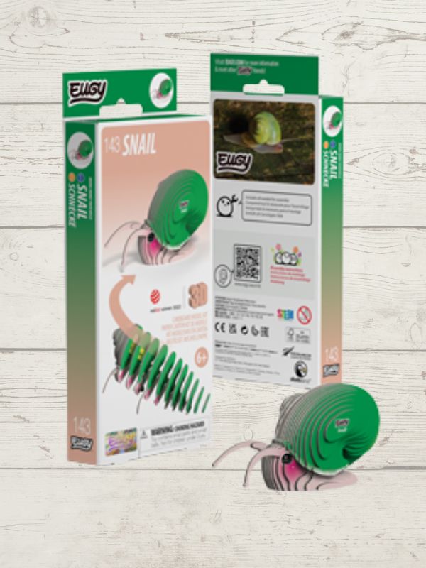 EUGY 143 Snail toy shown with its retail packaging on a light wood background. The product is a green coiled snail toy with pink and black striped legs, with two boxes visible behind it and one loose toy in the foreground.