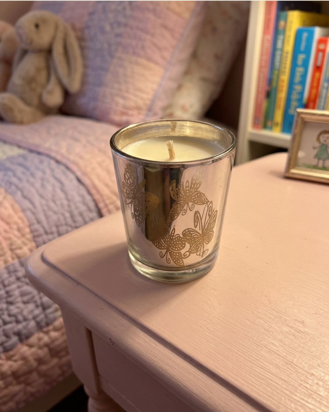 A lit scented candle in a clear glass jar with a gold floral pattern, sitting on a light pink bedside table in a cozy bedroom setting.
