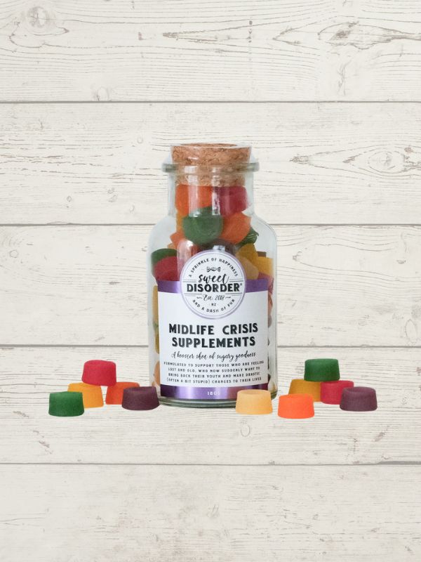 A clear glass bottle with a cork stopper and a white label reading 'Midlife Crisis Supplements' by Sweet Disorder, filled with colorful gummy candies, displayed on a white wood plank background with several gummies scattered in front.