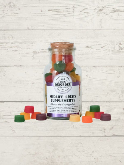 A clear glass bottle with a cork stopper and a white label reading 'Midlife Crisis Supplements' by Sweet Disorder, filled with colorful gummy candies, displayed on a white wood plank background with several gummies scattered in front.