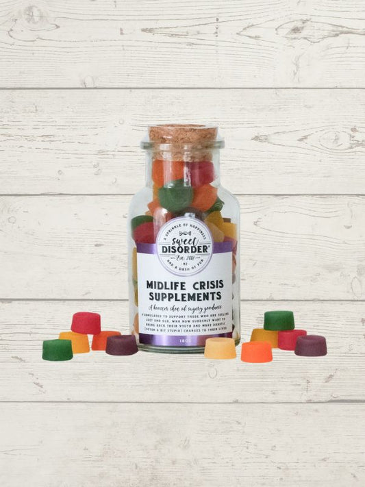 A clear glass bottle with a cork stopper and a white label reading 'Midlife Crisis Supplements' by Sweet Disorder, filled with colorful gummy candies, displayed on a white wood plank background with several gummies scattered in front.