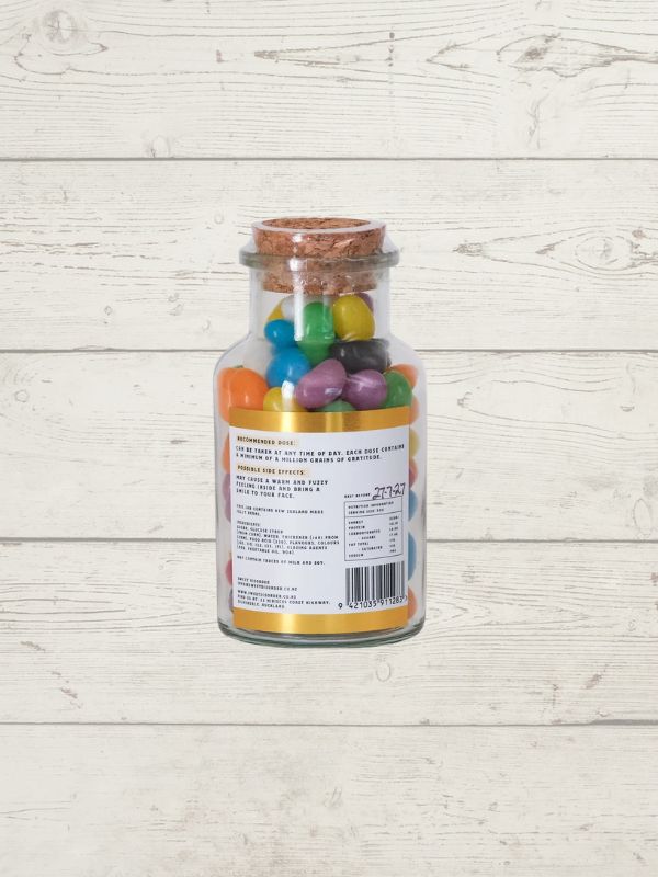 A clear glass bottle with a cork stopper filled with colorful jelly beans, shown against a light whitewashed wood plank background. The back of the bottle is labeled with text and a barcode.