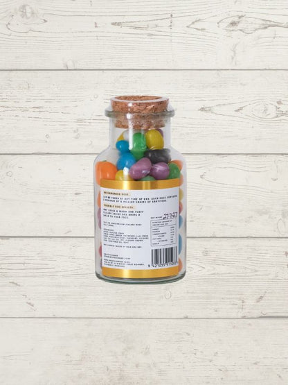 A clear glass bottle with a cork stopper filled with colorful jelly beans, shown against a light whitewashed wood plank background. The back of the bottle is labeled with text and a barcode.
