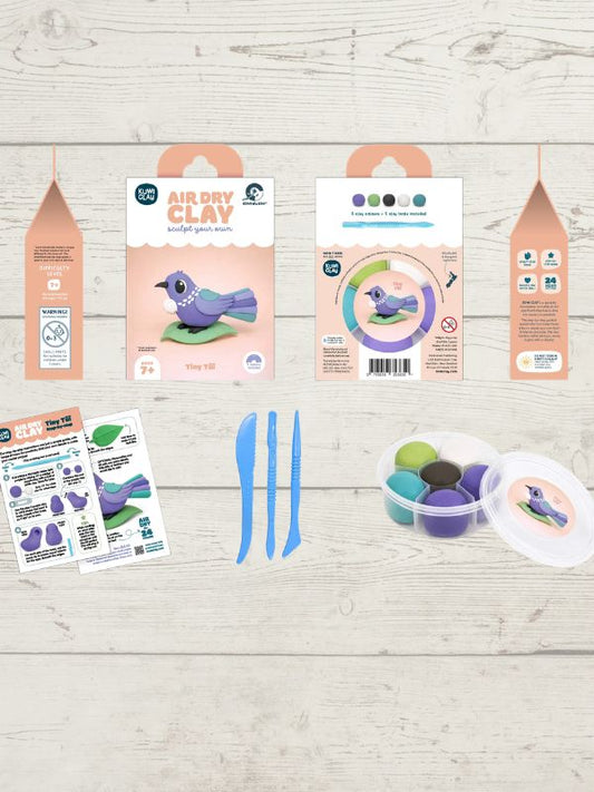 A clay art kit laid out on a light wood background, including two peach-colored hanging pouches, a package labeled Air Dry Clay, a printed instruction sheet, two blue sculpting tools, a round palette with multiple colored clay pieces, and a small printed card with a bird illustration.