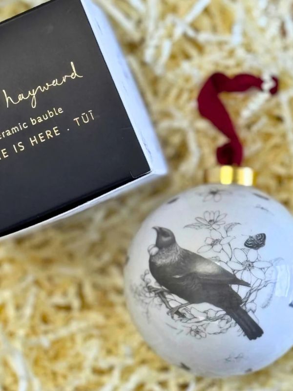 A white ceramic ornament featuring a black bird illustration and a red hanging ribbon, shown next to its black branded box on a bed of yellow paper shred.