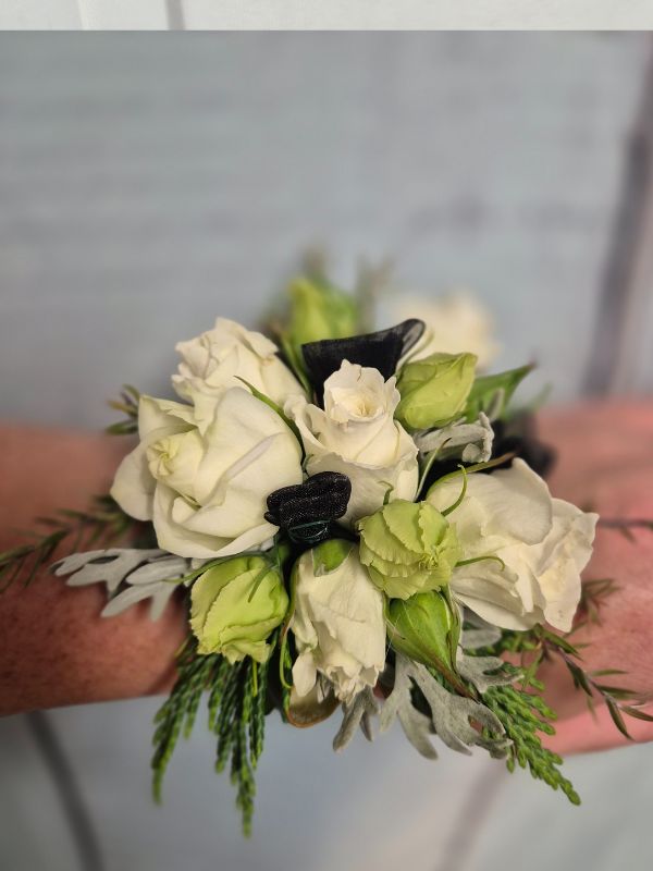 A wrist corsage featuring white and black flowers with green leaves, attached to a black organza ribbon.