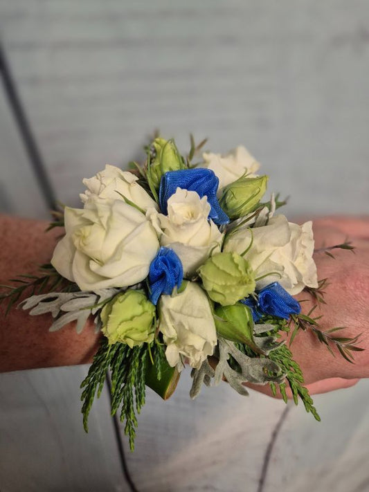 A wrist corsage featuring white and royal blue flowers with green leaves, attached to a royal blue organza ribbon.