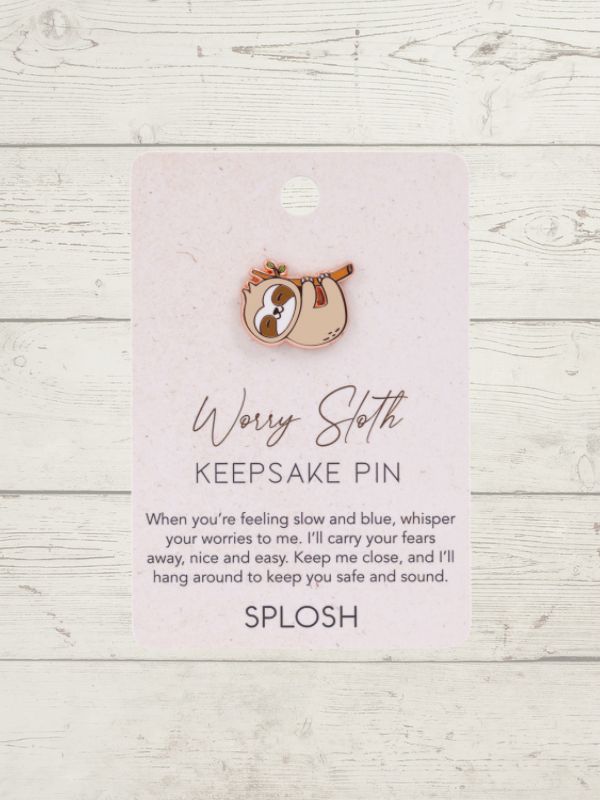 A small enamel keepsake pin shaped like a sloth resting on a pink card that reads 'Worry Sloth KEEPSAKE PIN' with a quote and the brand name 'SPLOSH', photographed against a whitewashed wood background.