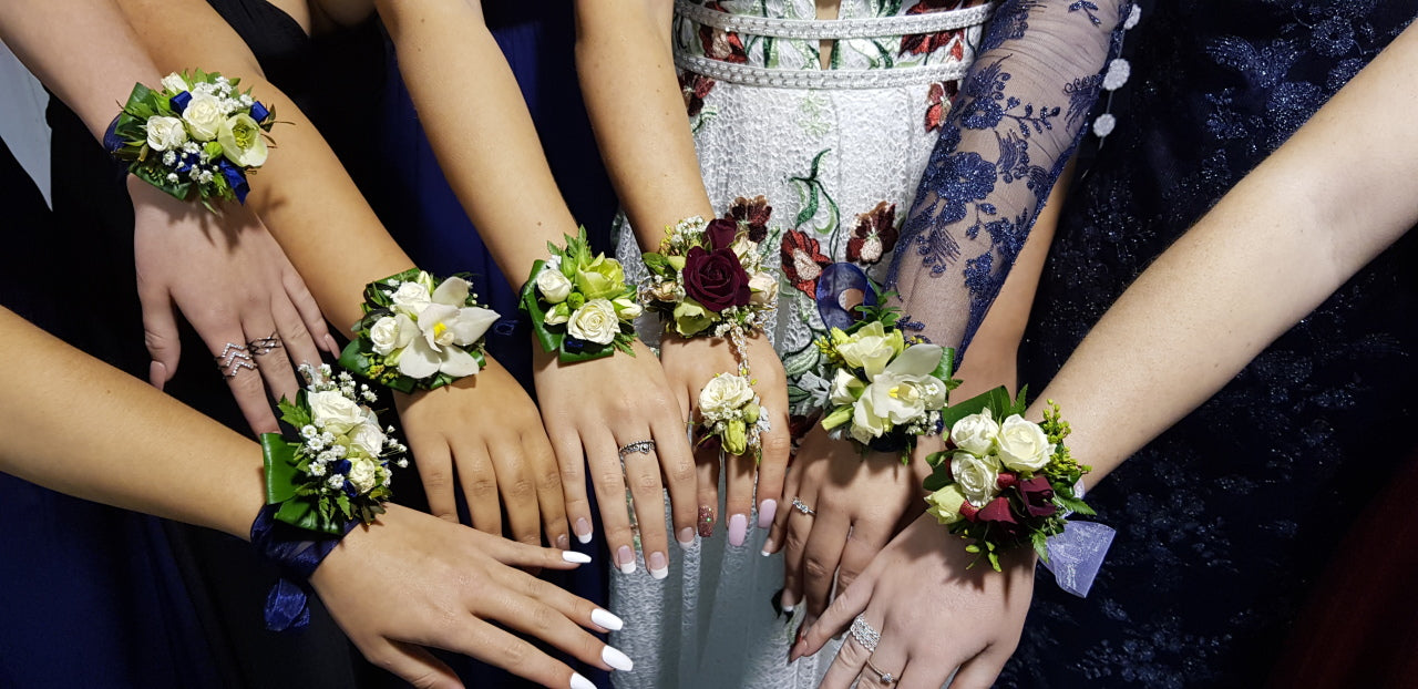 A group of bridesmaids' hands and wrists gathered together, each wearing a matching floral wrist corsage with white, green, and burgundy flowers. The background shows glimpses of wedding attire in navy blue, white, and lace.