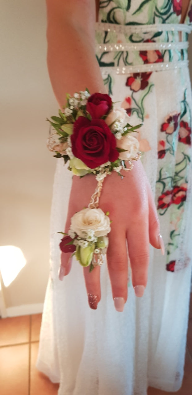 A close-up of a person's hands wearing a matching wrist and ring corsage set. The floral wrist corsage features a deep red rose, white carnations, and green leaves, while the ring corsage has white flowers and green leaves.