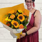A person holding a bouquet of flowers in yellows including sunflowers, chrysanthemums, gerberas wrapped in yellow and white paper with a yellow bow. 
