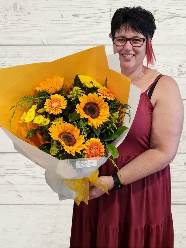 A person holding a bouquet of flowers in yellows including sunflowers, chrysanthemums, gerberas wrapped in yellow and white paper with a yellow bow. 