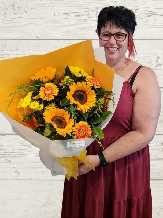 A person holding a bouquet of flowers in yellows including sunflowers, chrysanthemums, gerberas wrapped in yellow and white paper with a yellow bow. 